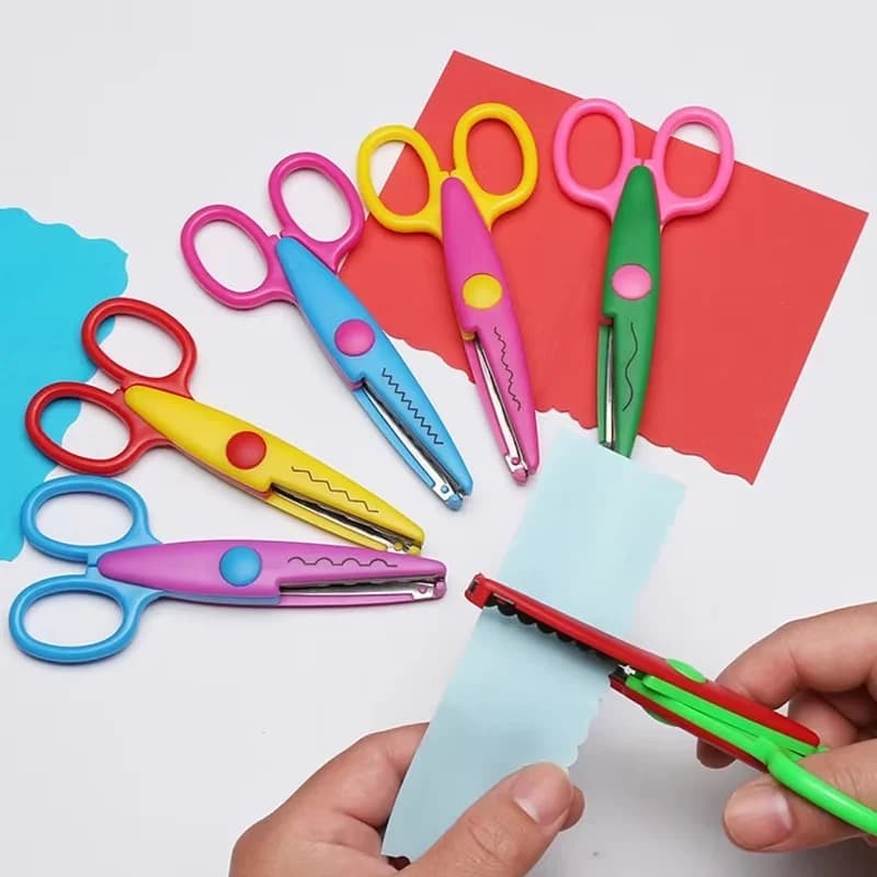 Six paires de ciseaux crantés aux branches colorées posées sur des papiers bleu et rouge, une main en utilise une pour découper un bord ondulé.