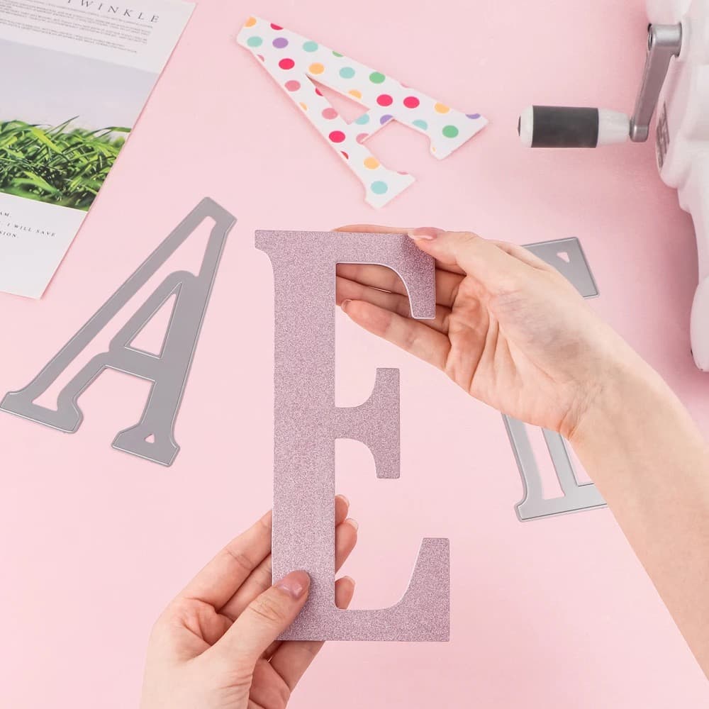 Grande lettre E découpée dans du papier pailleté rose tenue à deux mains, avec des matrices de lettres métalliques et un A à pois posés autour sur un fond rose.