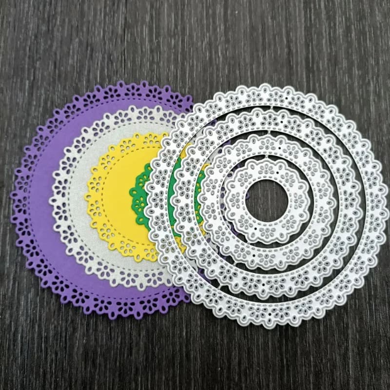 Matrices de découpe rondes à bordure dentelle emboîtées, posées sur les napperons de papier violet, gris et jaune qu'elles permettent de découper.