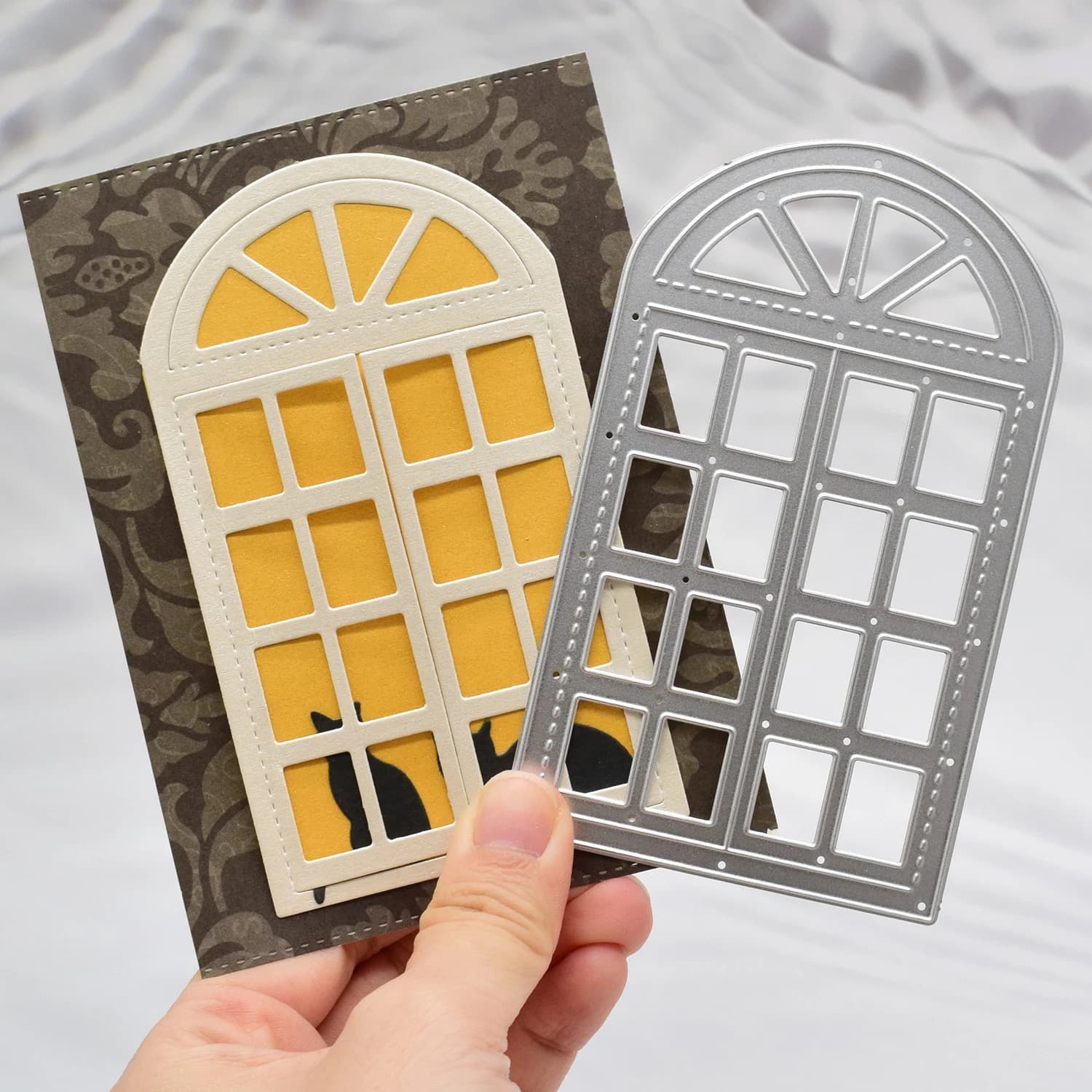 Matrice métallique de fenêtre cintrée tenue à la main devant la carte finie, où la fenêtre crème découpée laisse voir un fond jaune et une silhouette de chat.