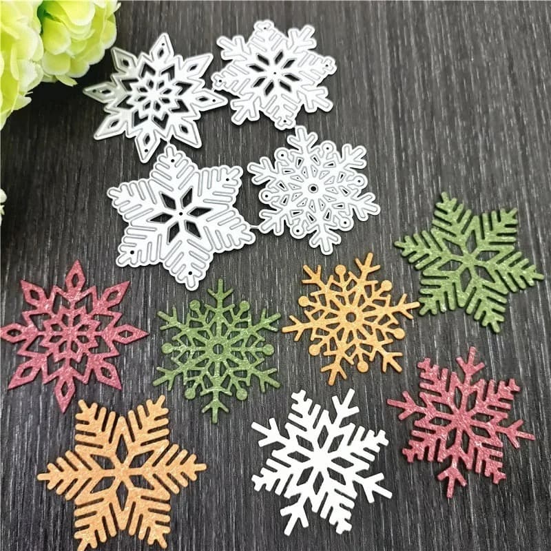 Matrices de découpe en forme de flocons de neige posées sur un plan de travail sombre, entourées de flocons découpés dans du papier pailleté de plusieurs couleurs.