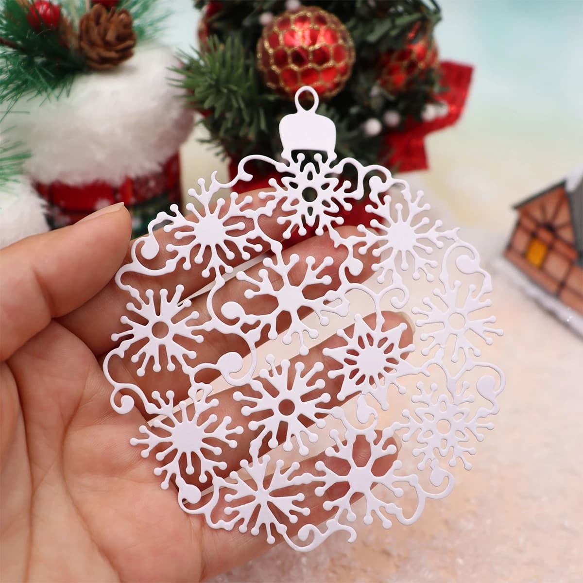 Boule de Noël en papier blanc entièrement ajourée de flocons, tenue entre deux doigts devant un décor de sapin et de boules rouges.