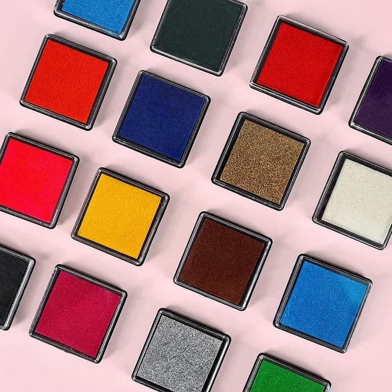 Petits encreurs carrés dans des couleurs variées, rouge, bleu, jaune, doré, argent et bordeaux, disposés en diagonale sur un fond rose pâle.