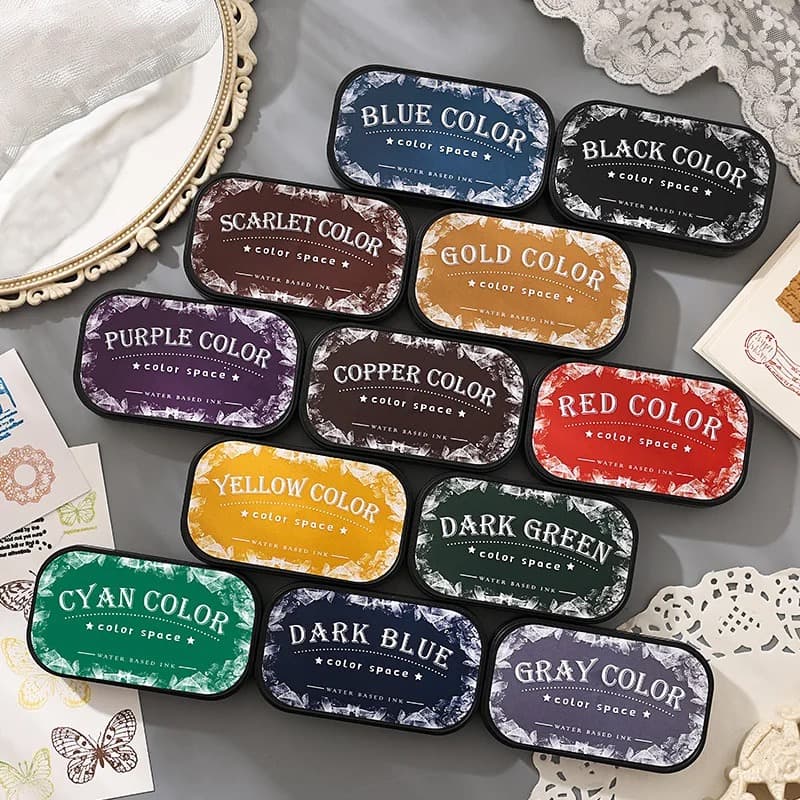 Douze encreurs ovales dont le couvercle porte le nom de la couleur, disposés sur un fond gris avec un miroir ancien, un napperon en dentelle et des cartes tamponnées.
