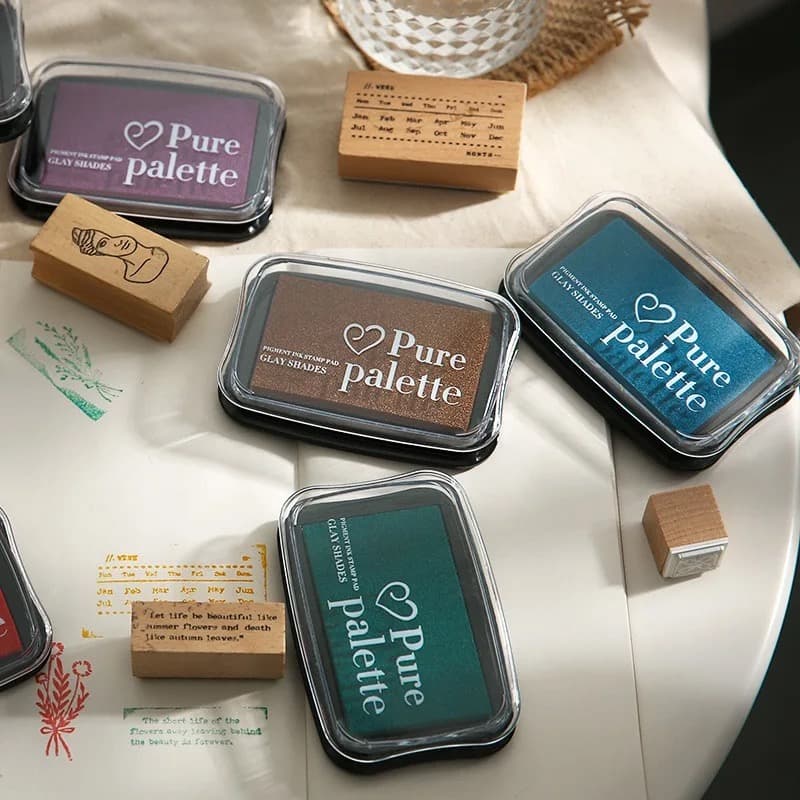 Plusieurs encreurs grand format dans des teintes prune, cuivre, bleu canard et vert sapin posés sur une table claire, avec des tampons en bois et des empreintes imprimées sur papier.