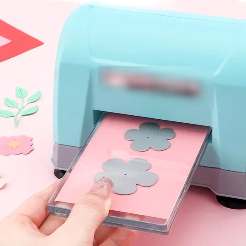 Machine de découpe manuelle turquoise à grand passage, une main engageant un sandwich de plaques avec une fleur découpée.