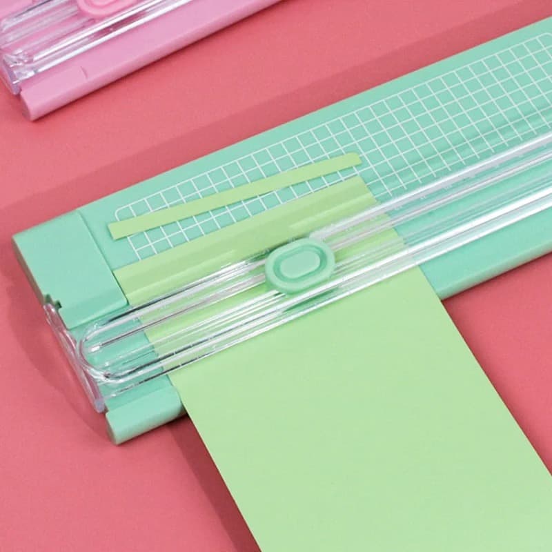 Massicot vert menthe avec rail transparent et plateau quadrillé, une feuille de papier vert engagée dessous, sur fond rose, un second massicot rose à l'arrière.