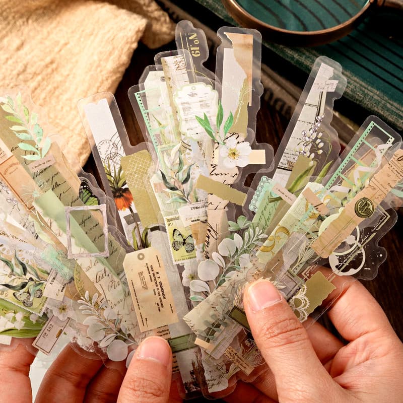 Bandes d'autocollants botaniques en teintes vertes et beiges, tenues en éventail entre deux mains.