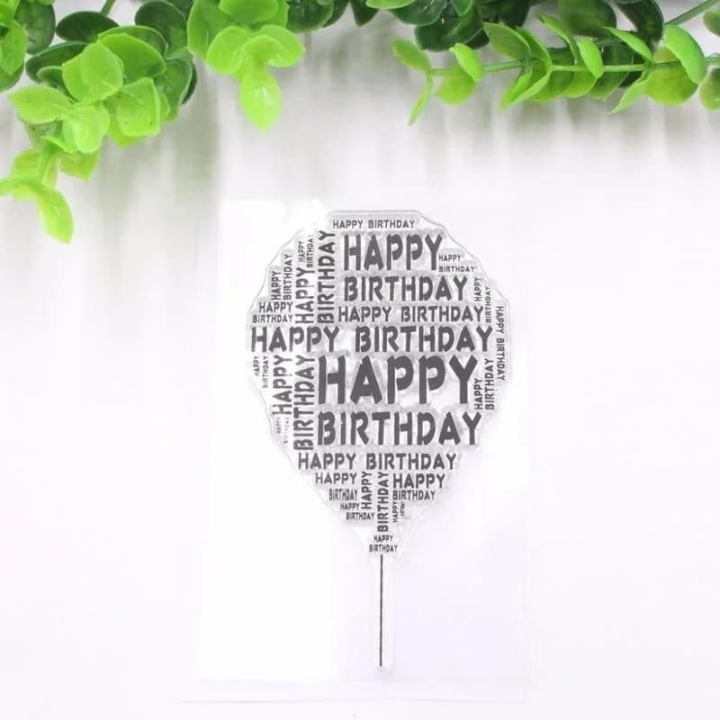 Tampon transparent en forme de ballon dont la silhouette est composée de la mention Happy Birthday répétée, posé sur fond blanc avec du feuillage vert.